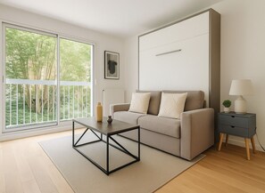 A modern living room with a sofa, a coffee table, a side table with a lamp, and a view of greenery through large windows. - Studio Élégant au 15eme (Paris)