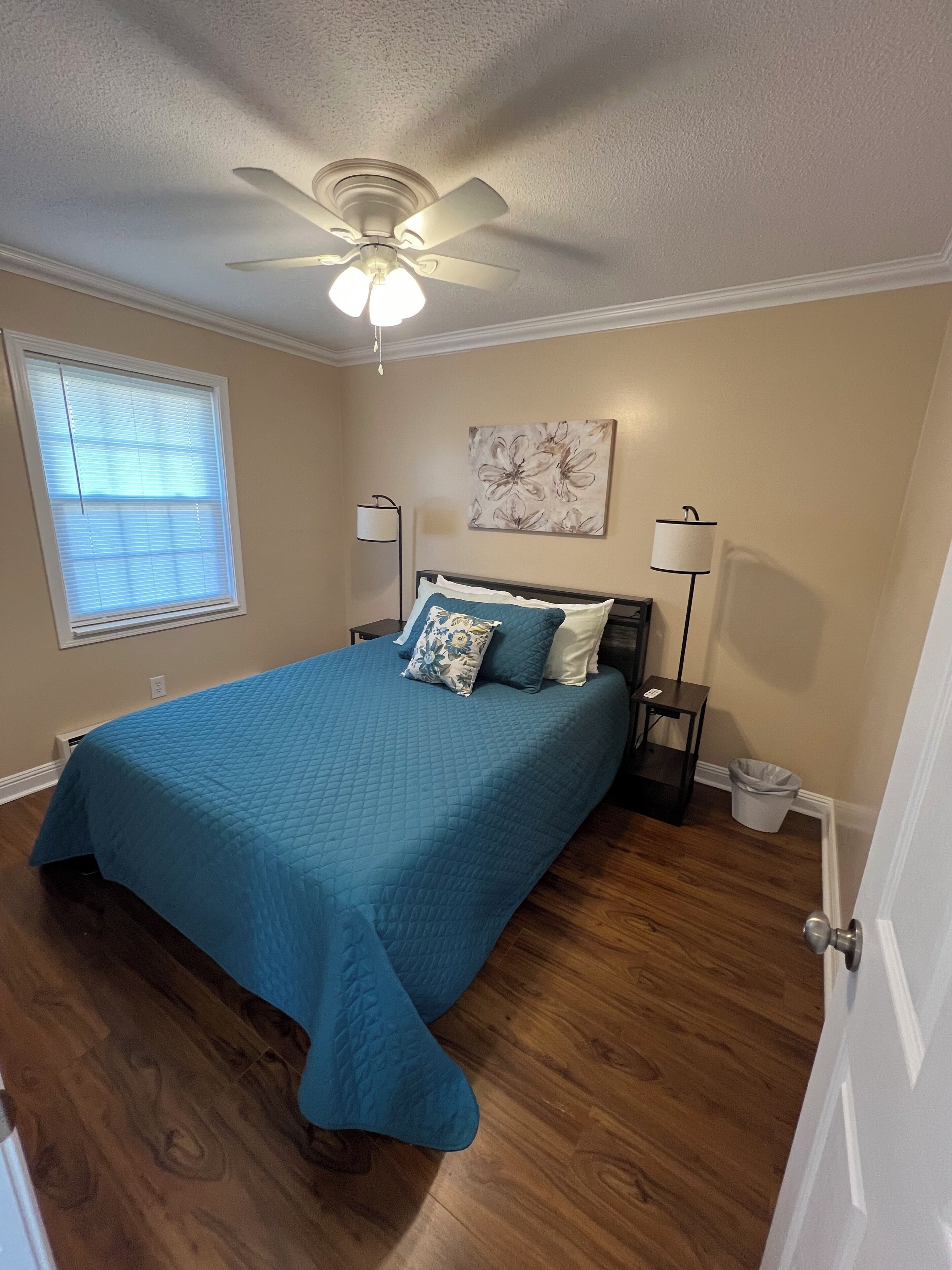 A bedroom with a bed, a ceiling fan, a window with blinds, a nightstand, a lamp, and a trash can.