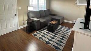Un salón con sofá gris, una mesa de centro, un televisor sobre un mueble bajo y una ventana con persianas.