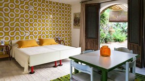 Habitación con cama, mesa y sillas. Se ve follaje verde a través de la ventana.