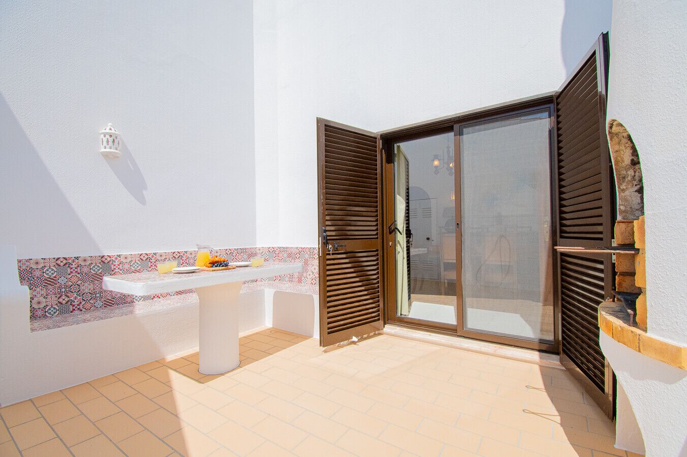 A sunlit patio with a white table set for breakfast, a patterned backsplash, and a sliding glass door with wooden shutters.