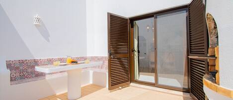 A sunlit patio with a white table set for breakfast, a patterned backsplash, and a sliding glass door with wooden shutters.