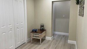 Un pasillo con suelo de madera, una puerta blanca, una pequeña mesa de madera con adornos y un espejo fijado a la pared.