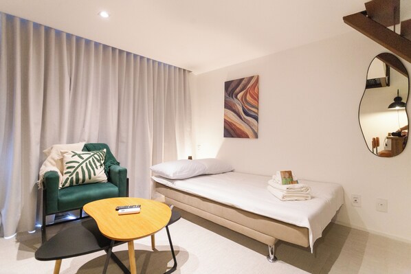 Habitación de hotel con un sillón verde, una pequeña mesa de madera, una cama, un espejo grande y un cuadro en la pared.