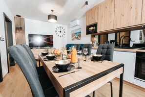 A modern kitchen with a wooden table set for dining, a clock on the wall, and a TV in the background.