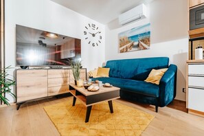 A living room with a blue sofa, a coffee table with baked goods, a clock, and a kitchen in the background.