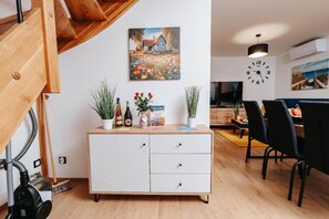 Un espacio habitable moderno con una escalera de madera, un comedor con sillas negras, un mueble blanco con plantas y botellas, y un reloj de pared.