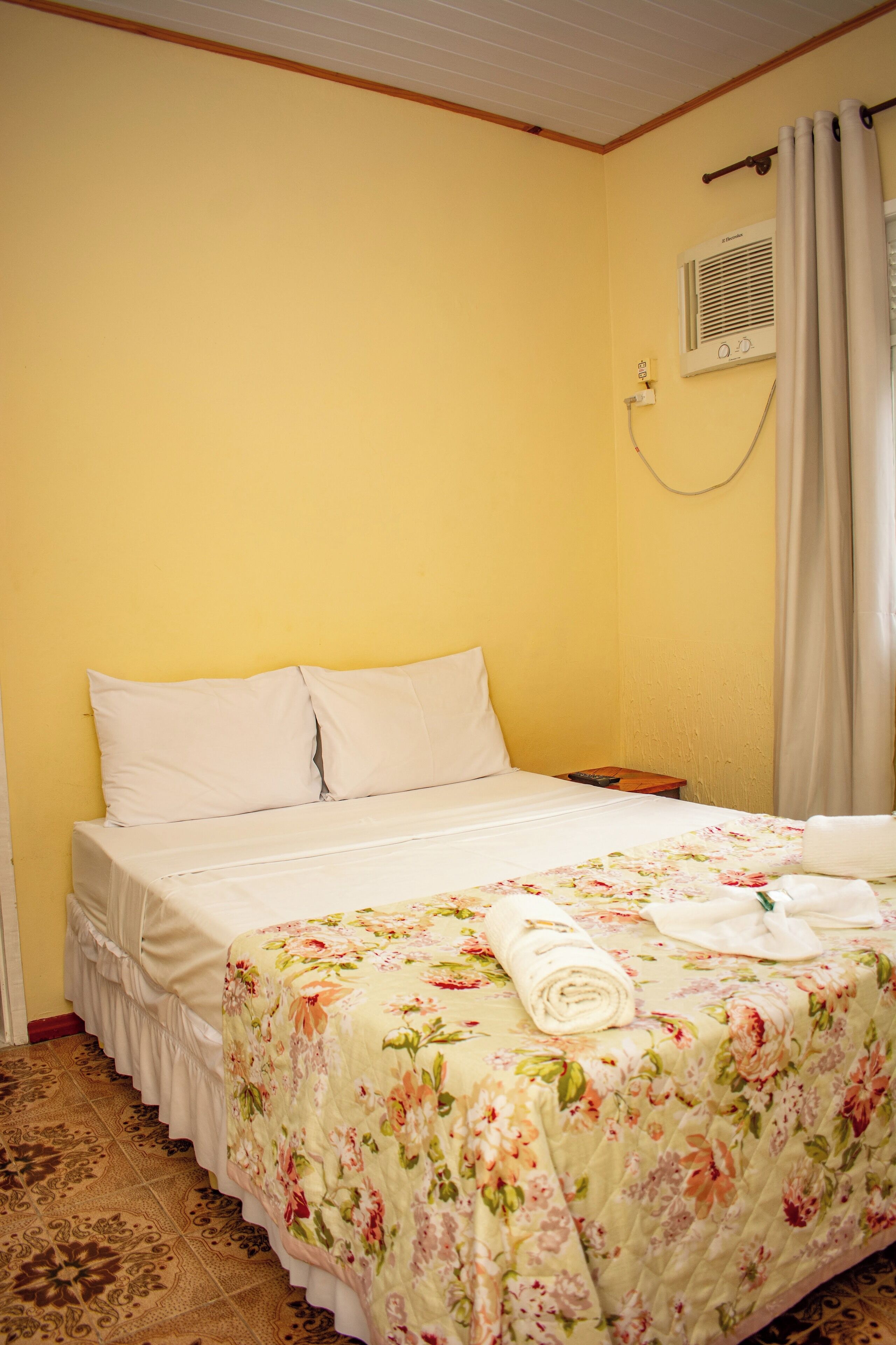 Una habitación de hotel con cama, cubrecama floral, almohada blanca, control remoto y aire acondicionado instalado en la pared.