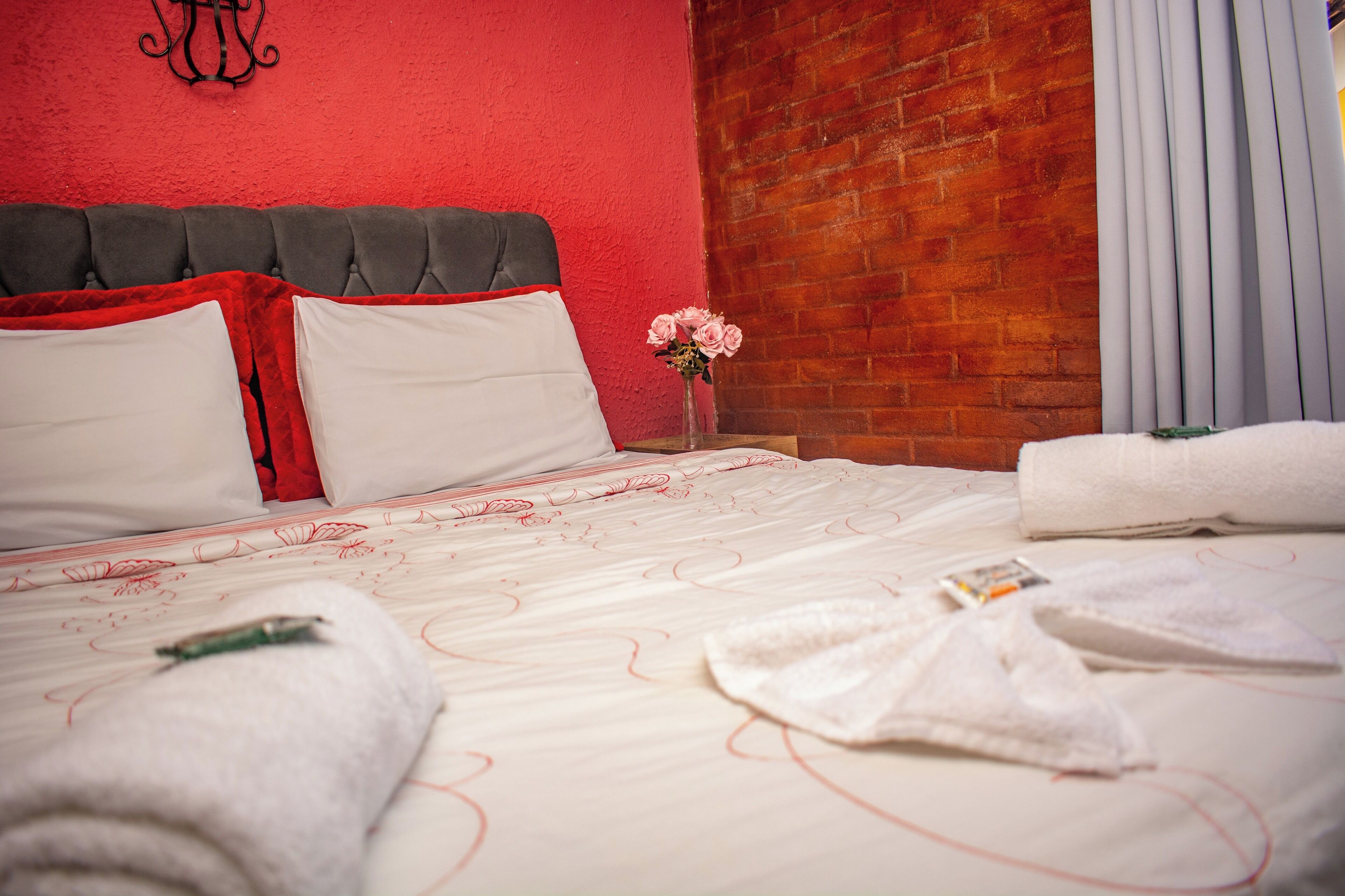 A neatly made bed with white and red bedding, a vase with pink flowers, and a brick wall in the background.