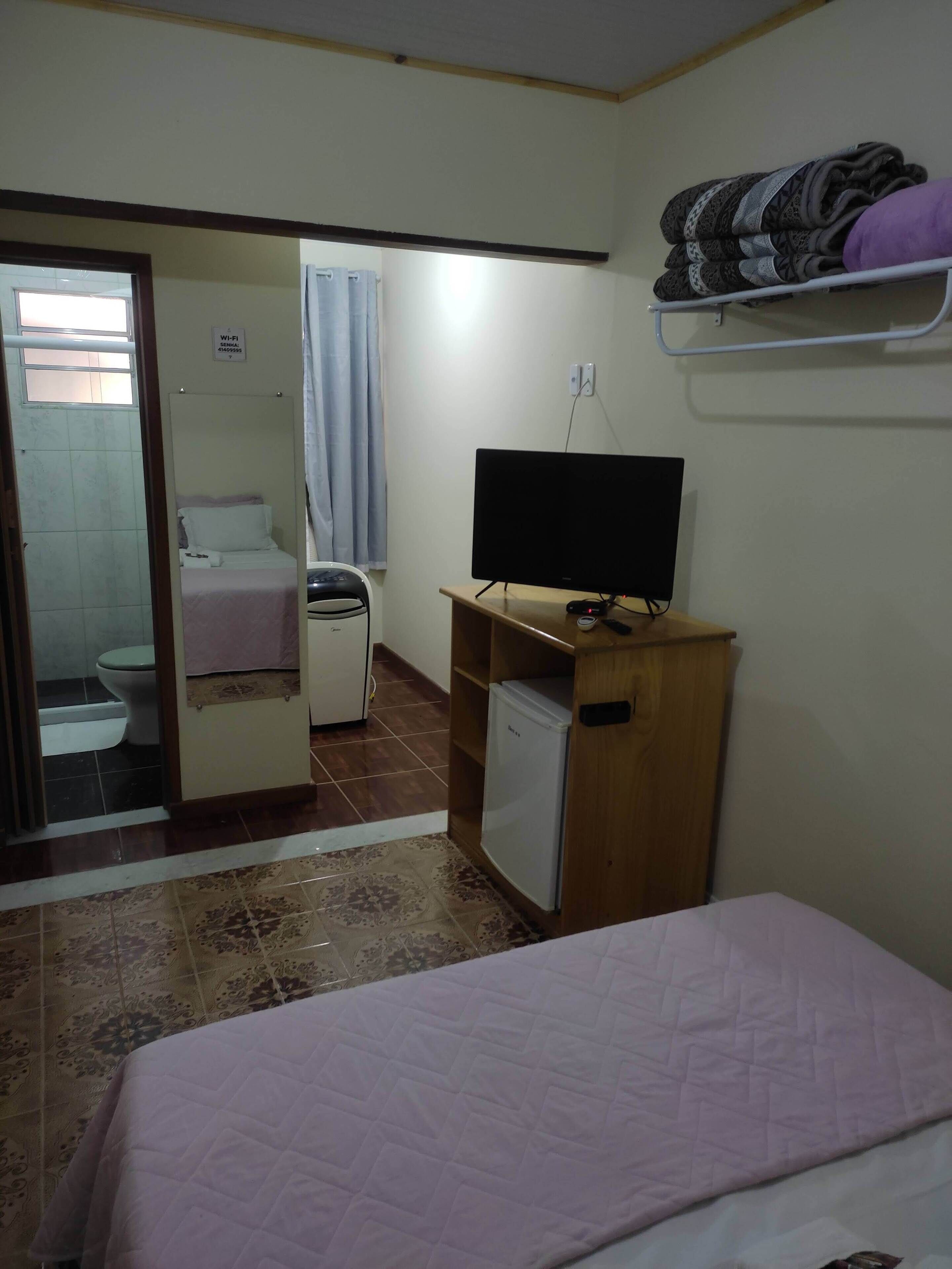 Un dormitorio con cama, escritorio con televisor, refrigerador pequeño y baño a la vista a través de una puerta abierta.