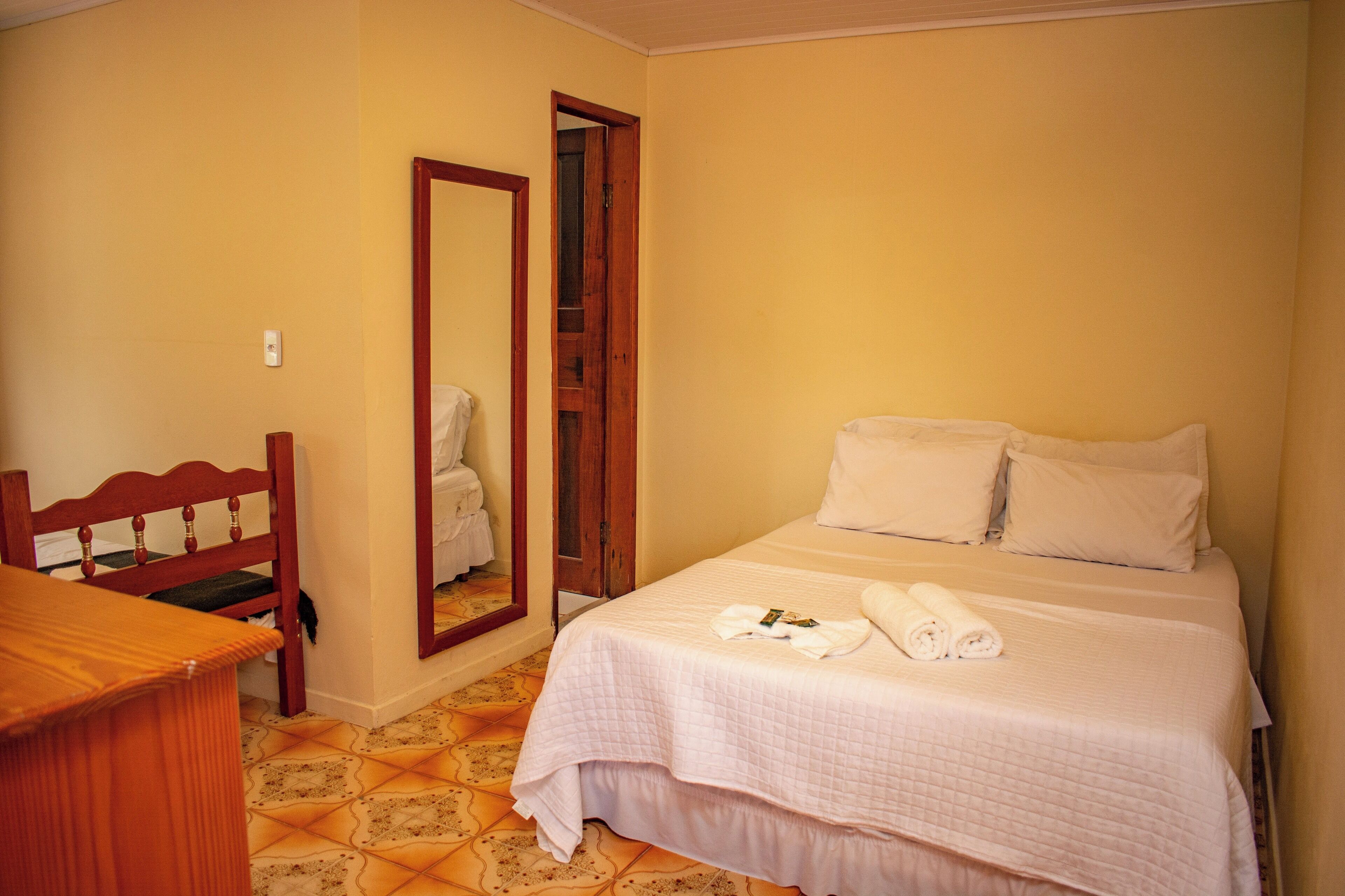 Una camera d'albergo con un letto, una sedia di legno, uno specchio e un pavimento piastrellato.