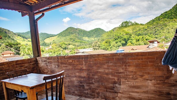 A wooden table and chairs on a balcony with a brick wall, overlooking a lush green valley and hills. - POUSADA KLEIN (Nova Friburgo)