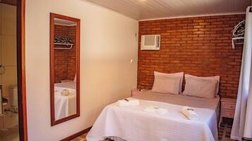 Quarto com parede de tijolos, cama com roupa de cama branca, mesa de cabeceira, espelho e janela com cortinas.