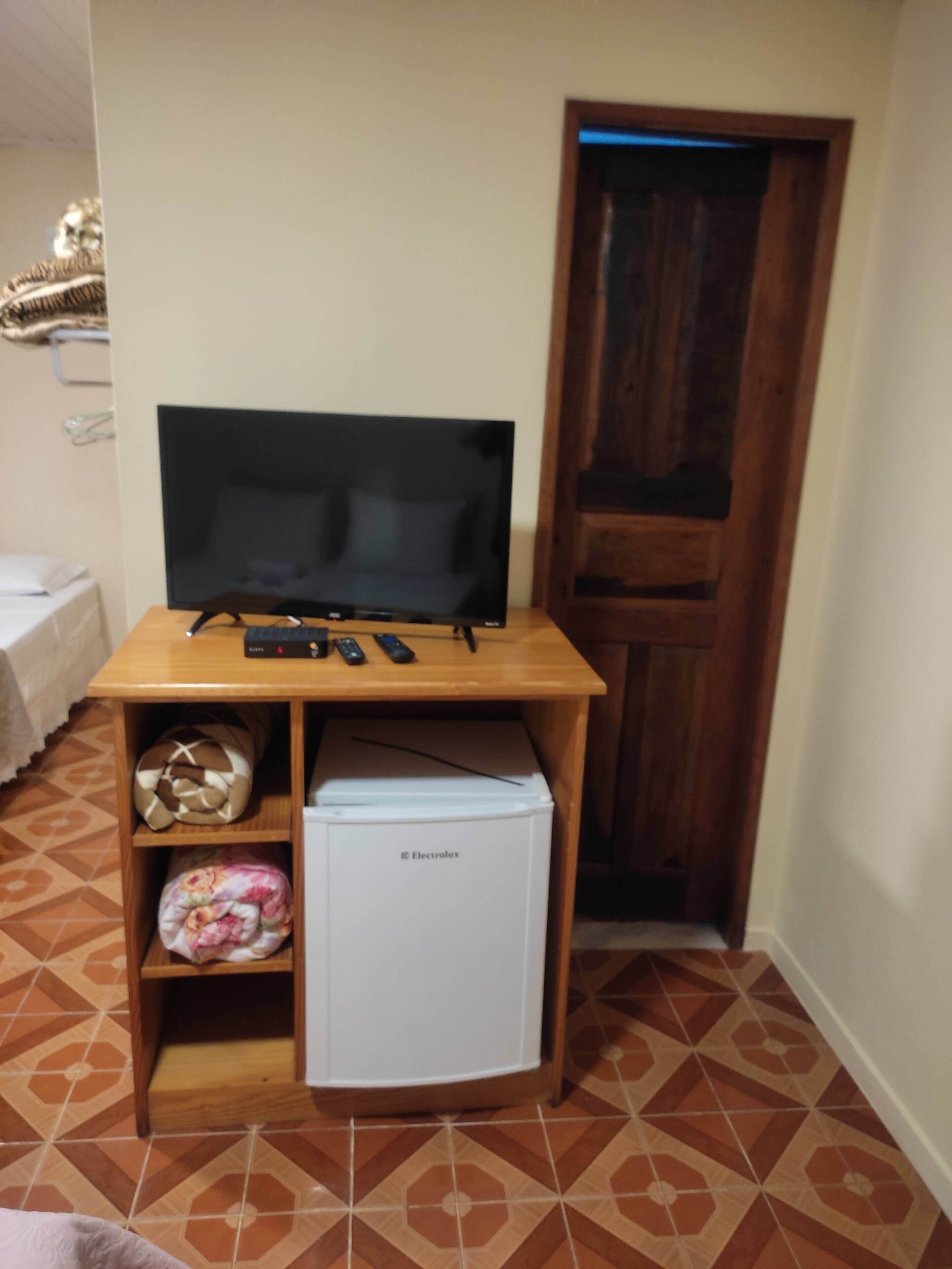 Una stanza compatta con un mobiletto in legno che sorregge una TV a schermo piatto, un piccolo frigorifero bianco e uno scaffale con un pallone da calcio e una coperta.
