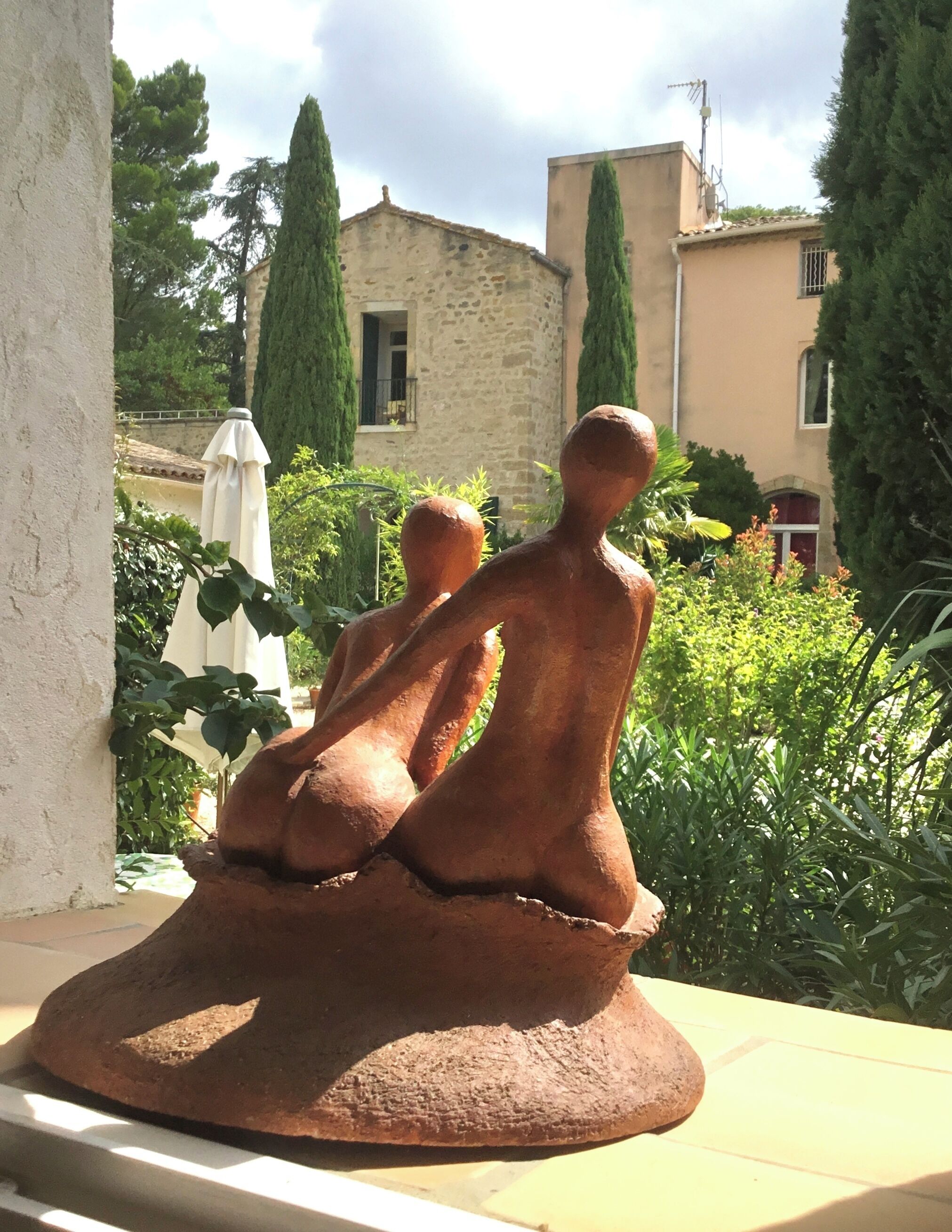 A sculpture of two figures seated on a pedestal in a garden setting with buildings and trees in the background.