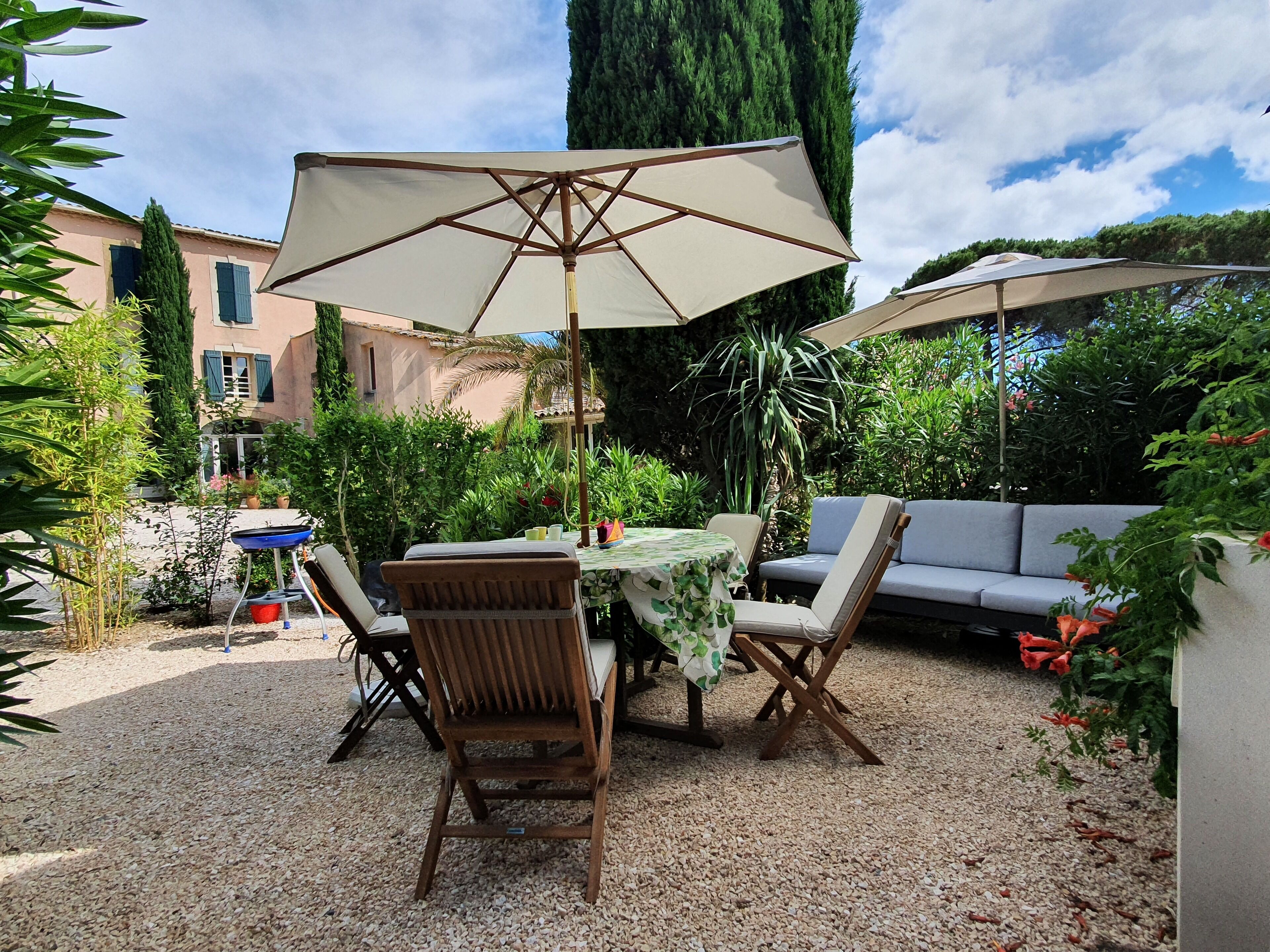 An outdoor patio with a table and chairs, surrounded by greenery and a building in the background.