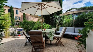 An outdoor patio with a table and chairs, surrounded by greenery and a building in the background.