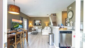 Un salón amplio con una mesa de comedor, sillas, un sofá y una mesa de centro. Se observa una cocina con lavavajillas y un reloj en la pared.