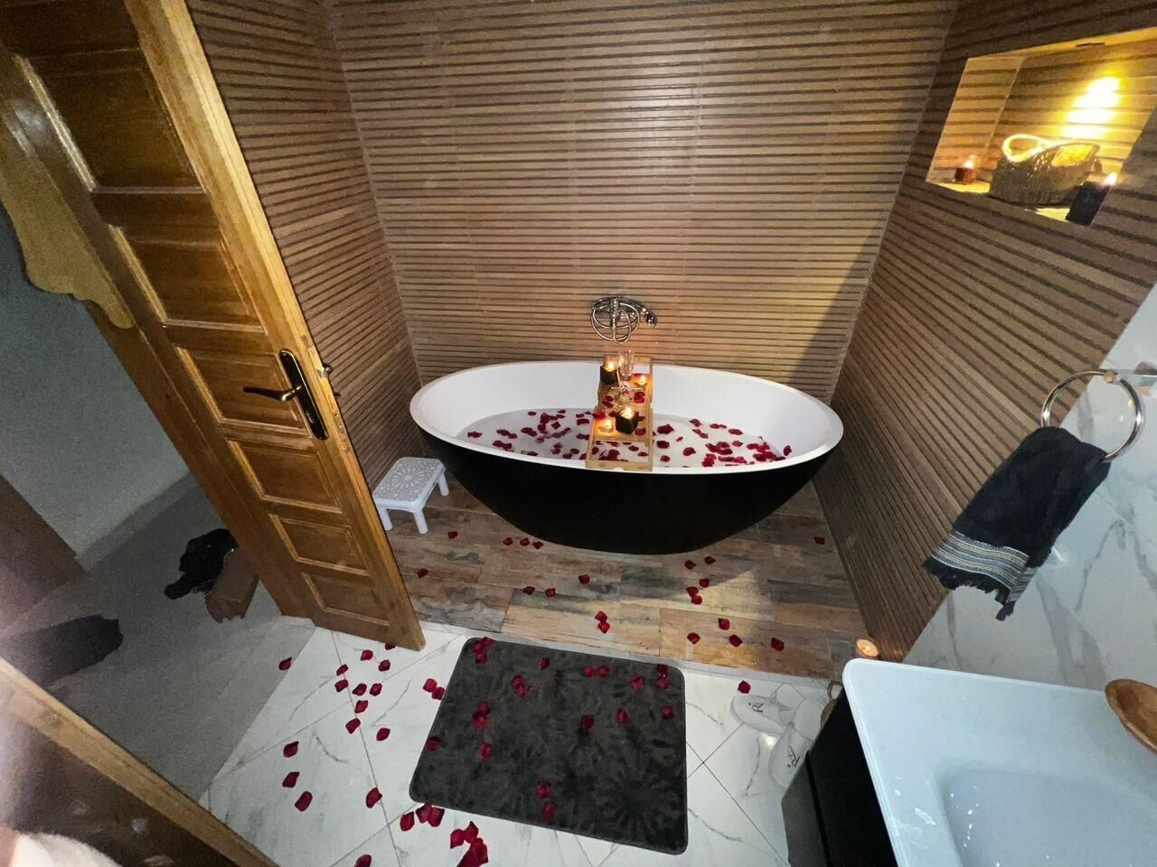 A bathroom with a freestanding bathtub, wooden panel walls, a small stool, and scattered rose petals on the floor.