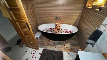 A bathroom with a freestanding bathtub, wooden panel walls, a small stool, and scattered rose petals on the floor.