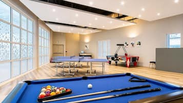 Une table de billard avec des billes et des queues, une table de ping-pong et une table de baby-foot dans une pièce bien éclairée, dotée de grandes fenêtres.