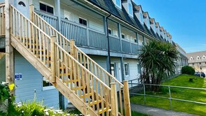 A row of coastal-style buildings with a wooden staircase leading to the entrance, surrounded by a grassy area with daisies. - Two Bedroom Apartment Easy Access (Ilfracombe)