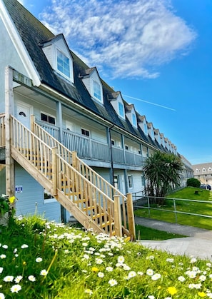 A row of coastal-style buildings with a wooden staircase leading to the entrance, surrounded by a grassy area with daisies.