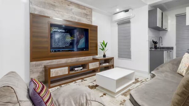 A modern living room with a flat-screen TV mounted on a wooden wall, a white coffee table, and a grey sofa.