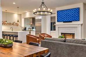 A modern living room with a fireplace, a dining area with a wooden table, and a kitchen in the background. - Premium Home Rooftop Pool Table 13 Beds (Nashville)