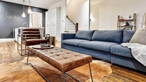 Un salon moderne avec un canapé bleu, un pouf en cuir et une table basse en bois. On aperçoit à l’arrière-plan un coin cuisine avec un meuble blanc et un luminaire suspendu.