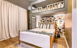 A bedroom with a bunk bed, a large window with curtains, a patterned wall, and a bedside table with a lamp.