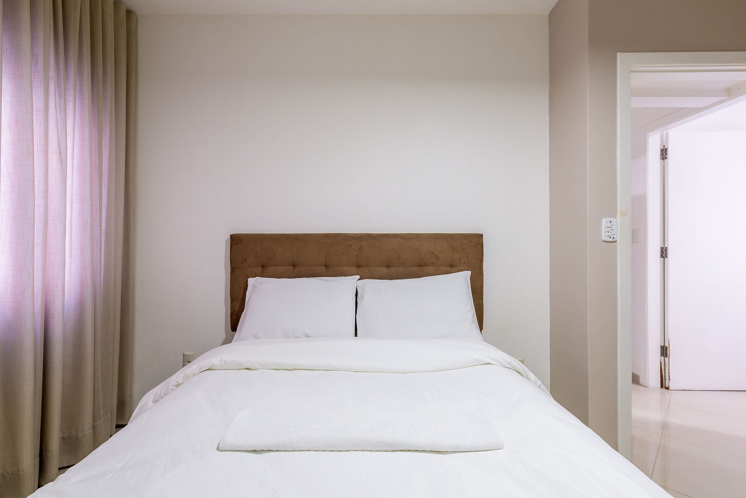 Een net opgemaakt bed met witte lakens in een kamer met een effen muur en een deur aan de rechterkant.