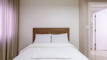 Una cama bien tendida con sábanas blancas en una habitación con pared lisa y una puerta a la derecha.