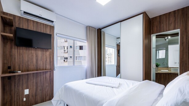 A modern hotel room with a flat-screen TV mounted on a wooden panel, a large mirror, and a neatly made bed. - Apt 4min From sea in BC Close to Everything Rta042 (Balneário Camboriú)
