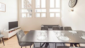 A dining area with a large table set for six, a television on a white cabinet, and a staircase visible through the windows.