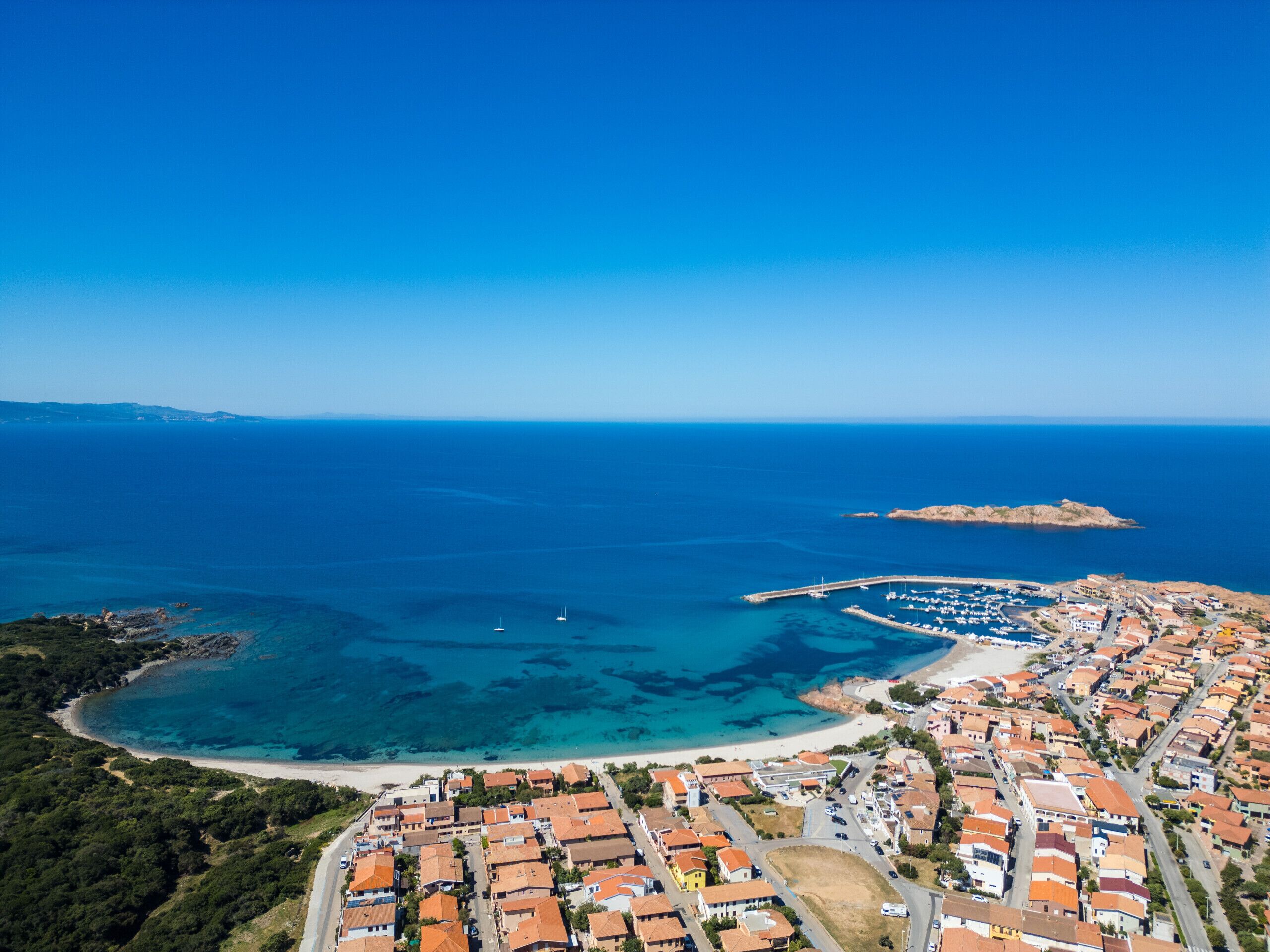 An aerial view of a coastal town with a marina, clear blue waters, and a beach.