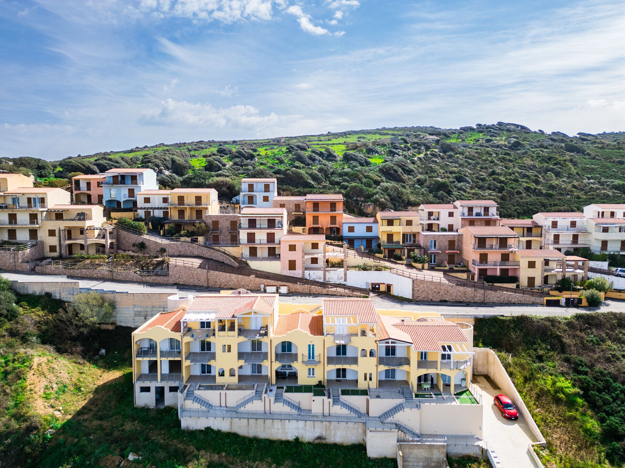 A coastal residential area with multi-story buildings, a prominent yellow building with balconies, and a red car on a narrow road.