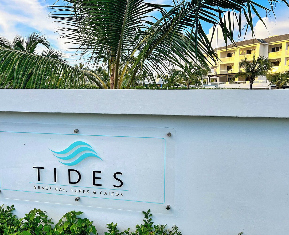 Ein Schild mit der Aufschrift 'TIDES' und einem Logo, vor einem Gebäude mit weißer Fassade und Palmen im Hintergrund.