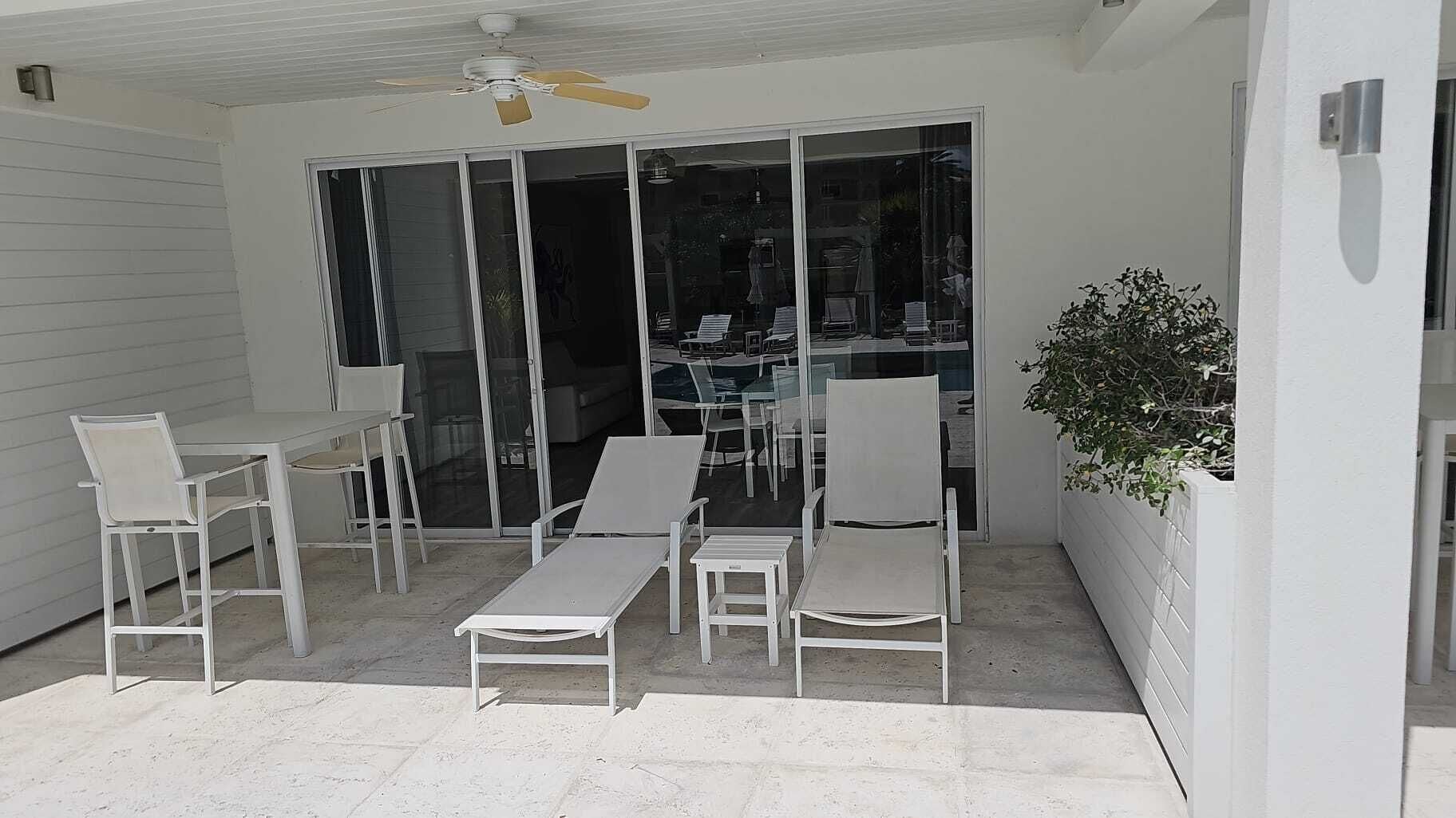 Un patio con mobiliario blanco, un ventilador de techo y un ventanal corredera que da a un espacio interior.