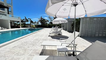 Een zwembadterras met witte ligbedden en parasols, een helderblauwe lucht en een zwembad.