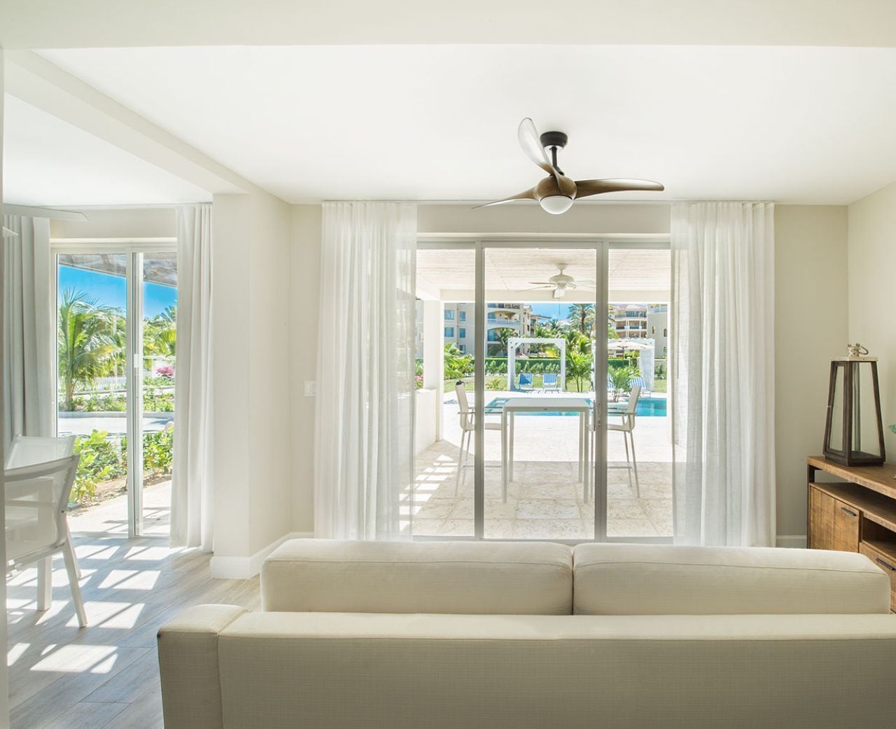 A modern hotel room with a large sliding glass door leading to a pool area, a ceiling fan, and a white sofa.