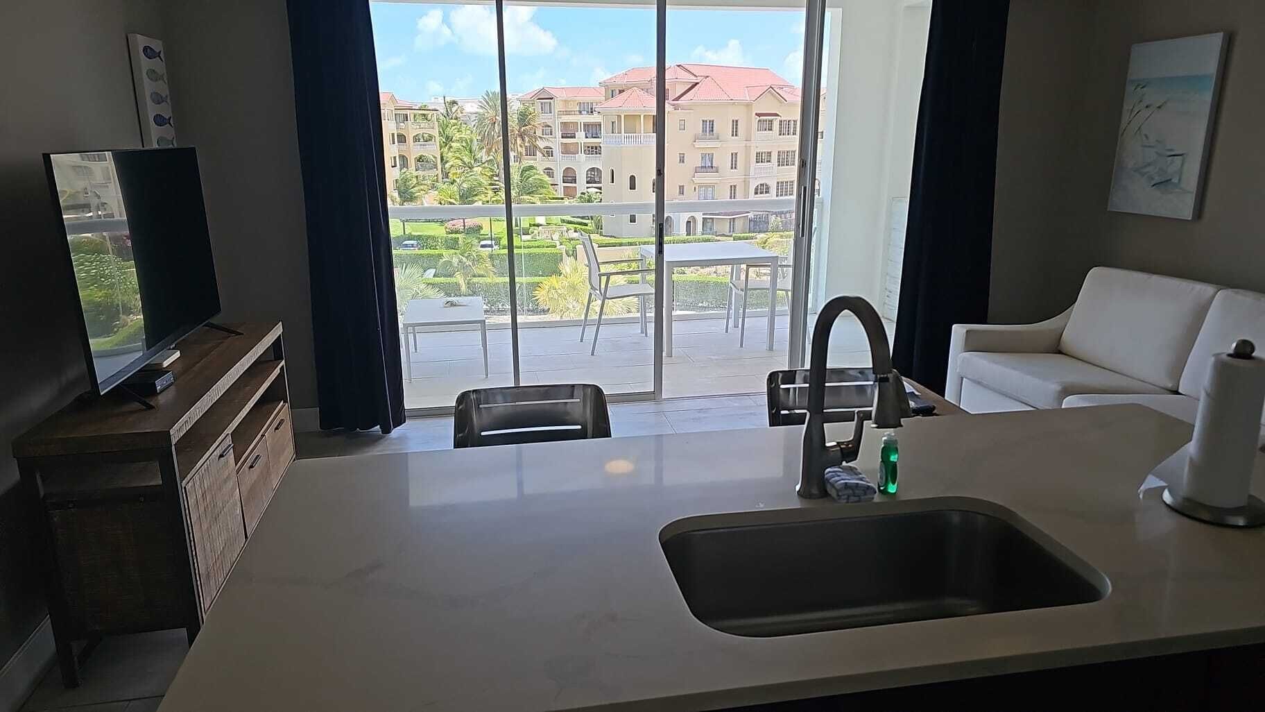 Une chambre d’hôtel moderne dotée d’une grande fenêtre, d’un canapé blanc, d’un coin cuisine avec un évier et d’un téléviseur à écran plat posé sur un meuble en bois.