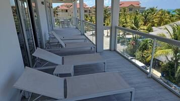 A balcony with white lounge chairs and a glass railing overlooking a residential area with palm trees and houses.