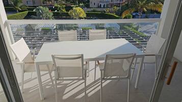 Un balcón con una mesa y sillas blancas, que ofrece vistas a un jardín cuidado y edificios.