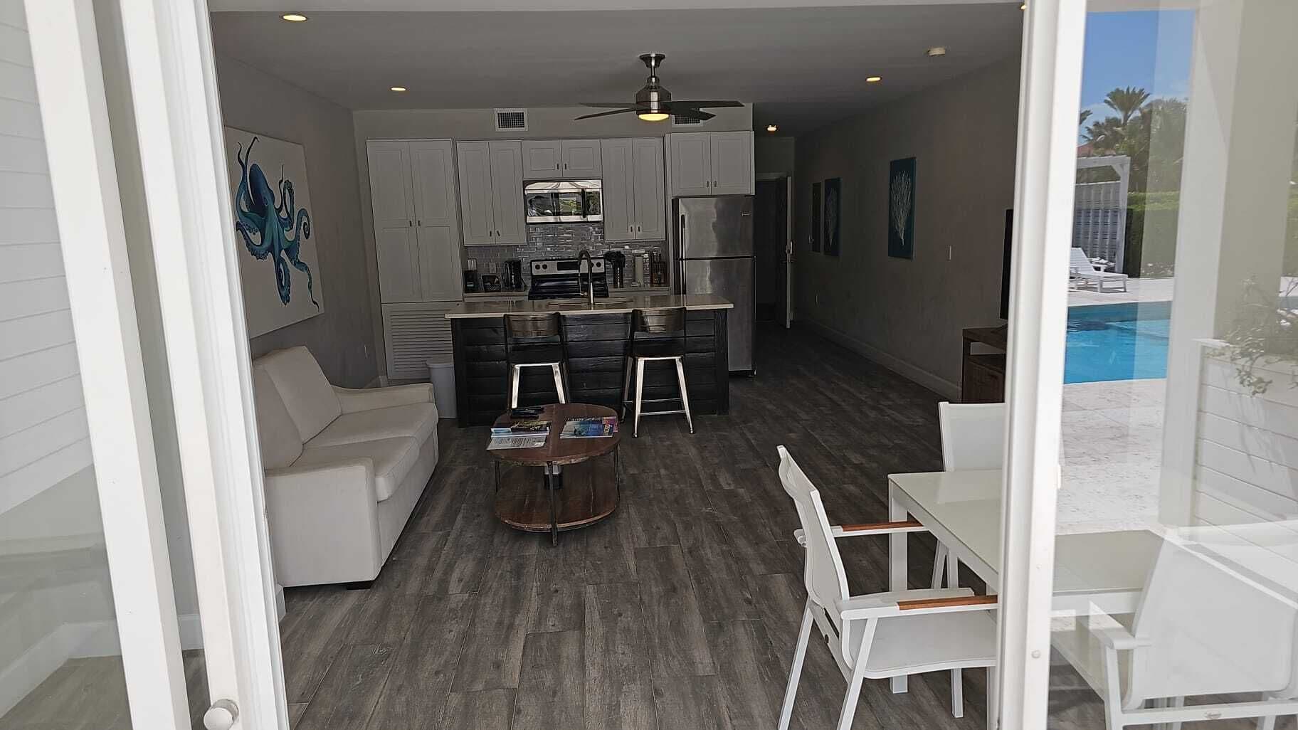 A modern kitchen with grey cabinets, a built-in oven, and a refrigerator. There is a dining area with a white table and chairs, and a small round table with books. A ceiling fan is visible above the kitchen.