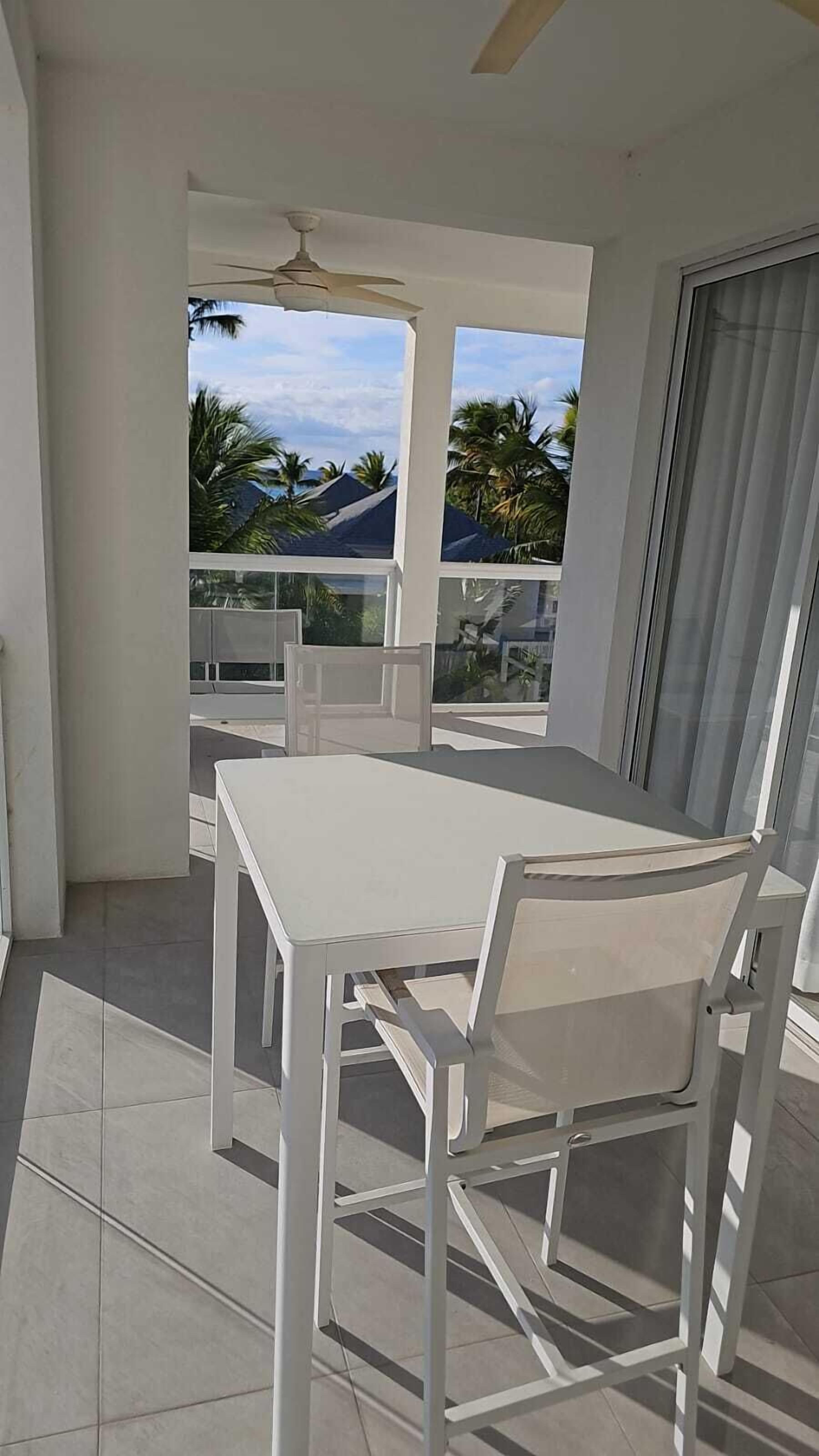Een balkon met een witte tafel en stoelen, een plafondventilator en uitzicht op palmbomen en gebouwen.