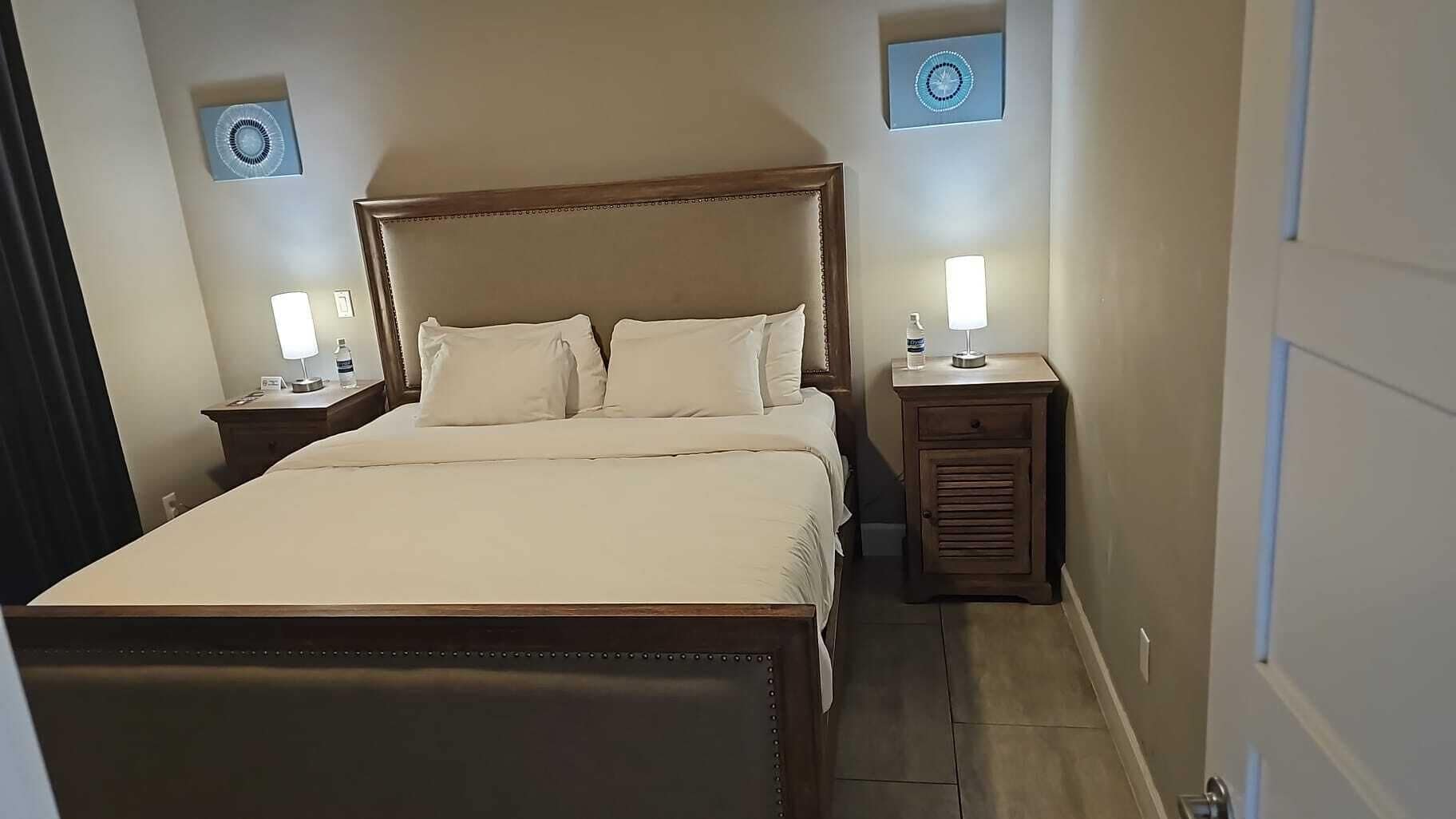 Een net opgemaakt bed met een houten hoofdbord, twee nachtkastjes met lampen, en een klein bijzettafeltje met een lamp.