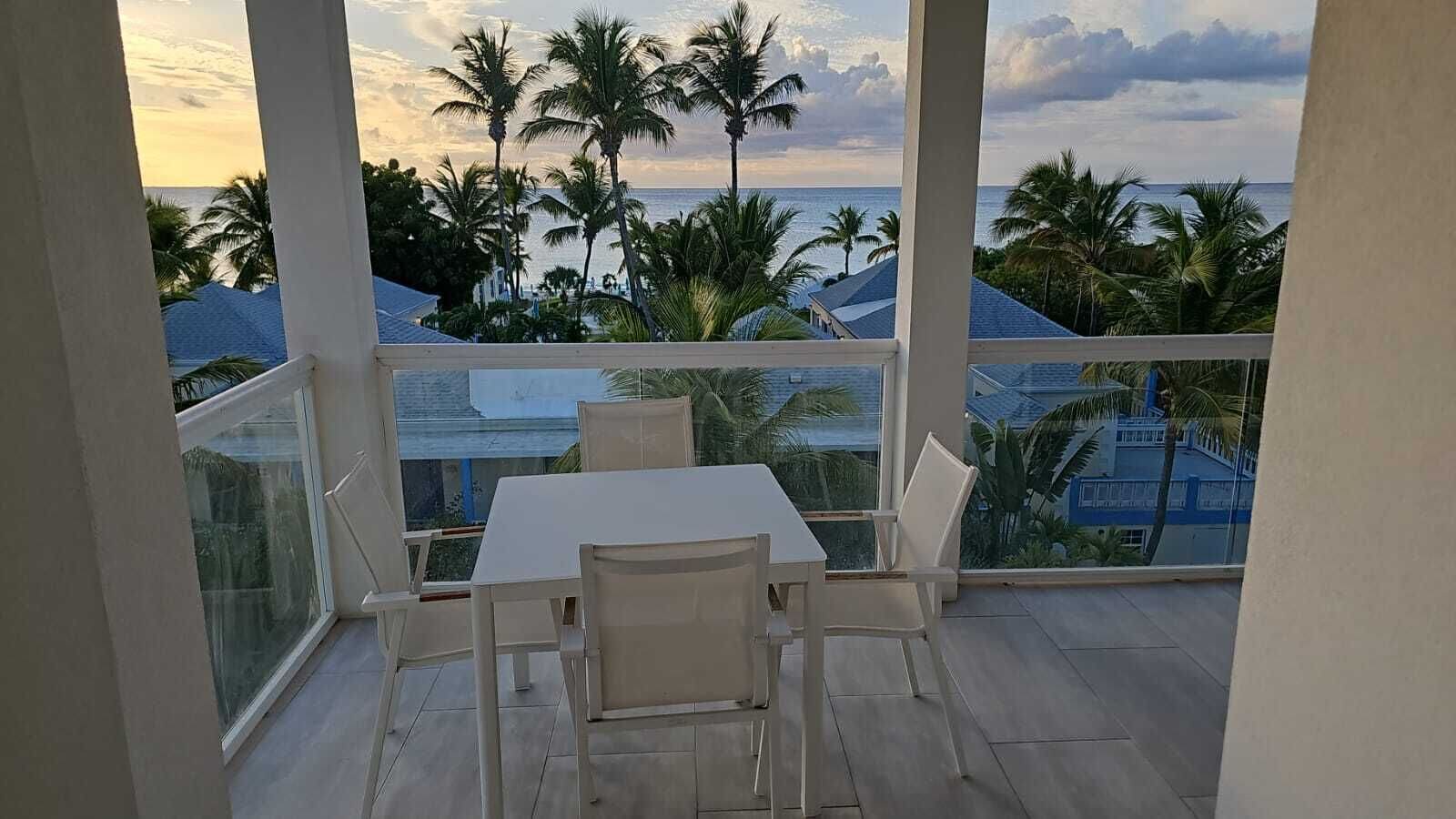 Un balcón con mesa y sillas, con vista a una playa con palmeras y al océano.
