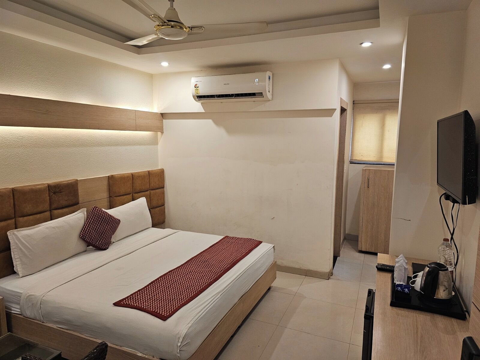 A hotel room with a bed, a ceiling fan, an air conditioning unit, a television, and a small kitchenette area.