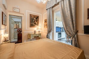 A hotel room with a bed, a desk with a lamp, a painting, and a view of a kitchen and dining area.